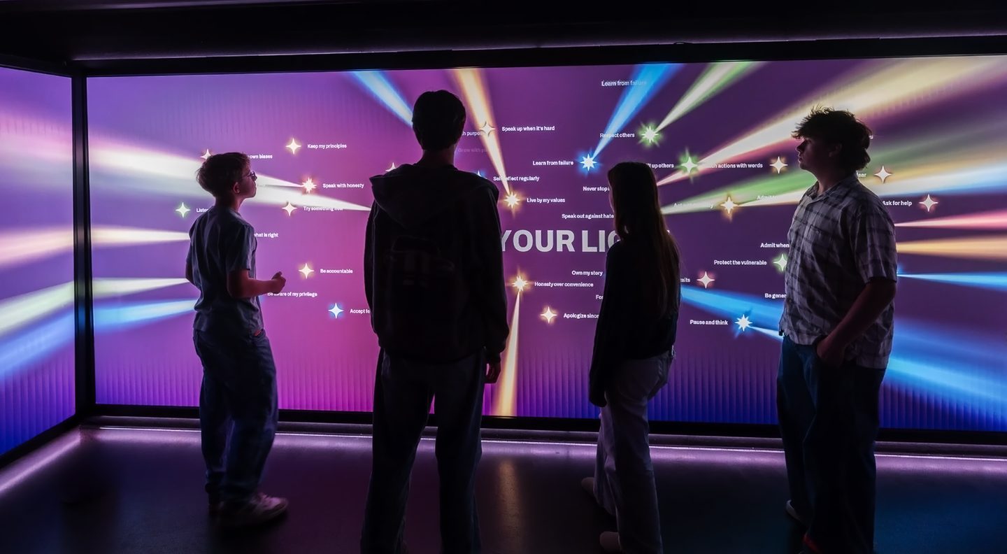 Visitors stand in a dark room facing an LED wall filled with beams of colored light and the words Share Your Light.